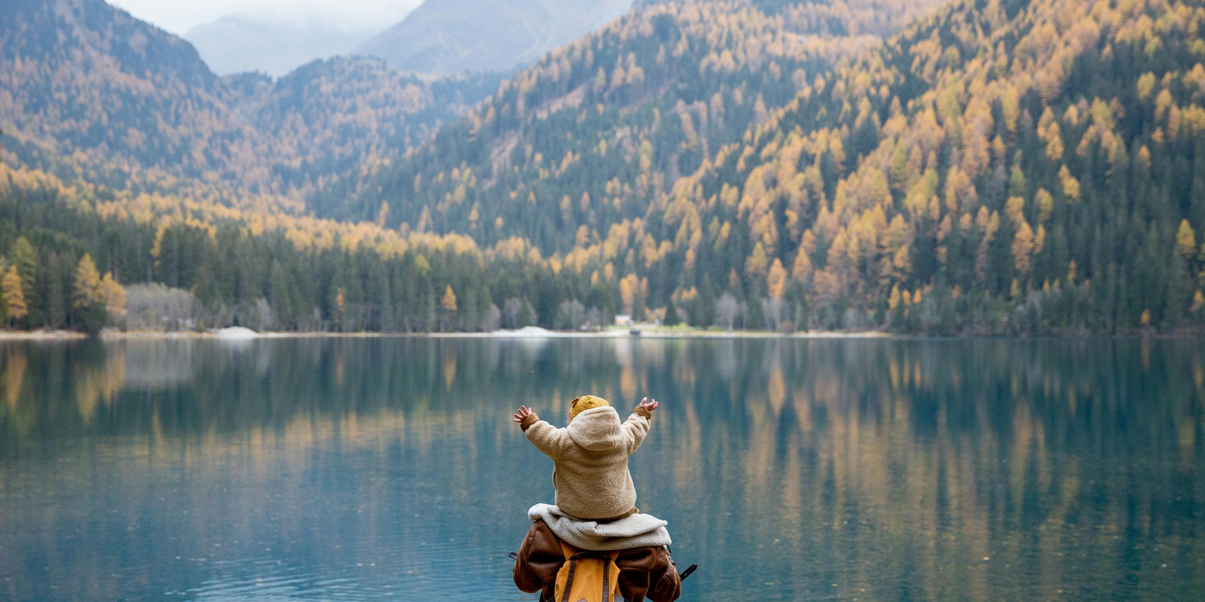 Ein Mann mit seinem Kind auf den Schultern steht vor einem See, umgeben von Bäumen. Das Bild verkörpert soziale Verantwortung und die Bedeutung von Familienbindung und Gemeinschaft.